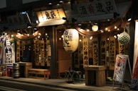 A Japanese restaurant with traditional wooden signage and hanging lanterns, illuminated warmly at night. The exterior includes wooden benches and barrels, with a chalkboard menu at the entrance. Decorative lights are strung along the storefront, adding to the inviting ambiance.