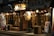 A Japanese restaurant with traditional wooden signage and hanging lanterns, illuminated warmly at night. The exterior includes wooden benches and barrels, with a chalkboard menu at the entrance. Decorative lights are strung along the storefront, adding to the inviting ambiance.