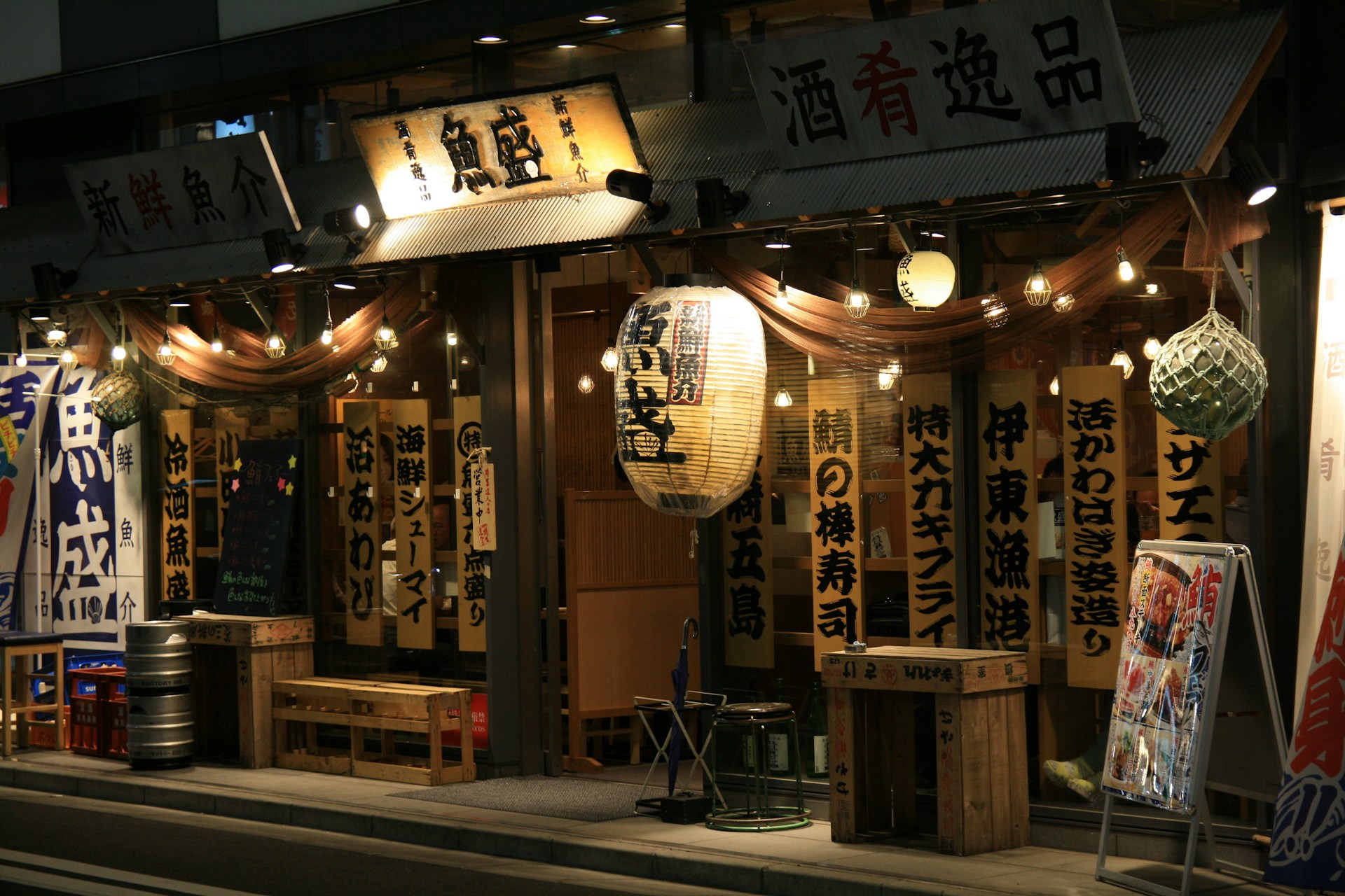 A Japanese restaurant with traditional wooden signage and hanging lanterns, illuminated warmly at night. The exterior includes wooden benches and barrels, with a chalkboard menu at the entrance. Decorative lights are strung along the storefront, adding to the inviting ambiance.