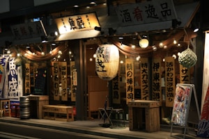 A Japanese restaurant with traditional wooden signage and hanging lanterns, illuminated warmly at night. The exterior includes wooden benches and barrels, with a chalkboard menu at the entrance. Decorative lights are strung along the storefront, adding to the inviting ambiance.
