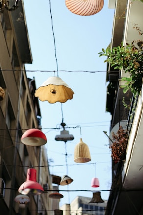 A collection of various types and colors of lampshades hanging on wires between two buildings. The sky is blue and visible in the background. The buildings have plants growing on them, adding some greenery to the scene. The lampshades are of different shapes such as circular, conical, and cylindrical, and are arranged at different heights.