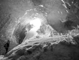 A person with a backpack is exploring a large ice cave. Sunlight filters through an opening, illuminating the smooth, textured ice walls and floor covered in snow.