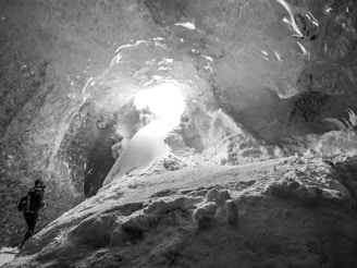 A person with a backpack is exploring a large ice cave. Sunlight filters through an opening, illuminating the smooth, textured ice walls and floor covered in snow.