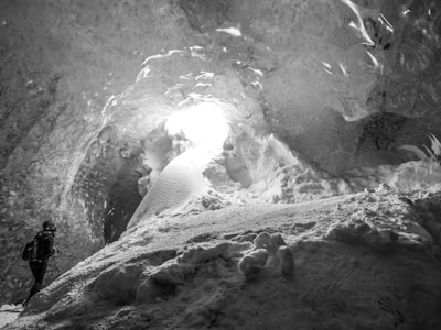 A person with a backpack is exploring a large ice cave. Sunlight filters through an opening, illuminating the smooth, textured ice walls and floor covered in snow.
