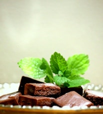 Several pieces of chocolate fudge are garnished with fresh green mint leaves. The chocolates are laid out on a surface, and the mint leaves add a vibrant touch to the arrangement.