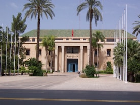 A large, stately building with a beige facade and a green tiled roof is surrounded by tall palm trees. A flagpole with a flag flying and several white flagpoles without flags are visible. The entrance is marked by large columns and a blue door. The surrounding area is landscaped with a variety of green plants and trees.
