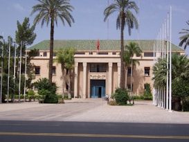 A large, stately building with a beige facade and a green tiled roof is surrounded by tall palm trees. A flagpole with a flag flying and several white flagpoles without flags are visible. The entrance is marked by large columns and a blue door. The surrounding area is landscaped with a variety of green plants and trees.