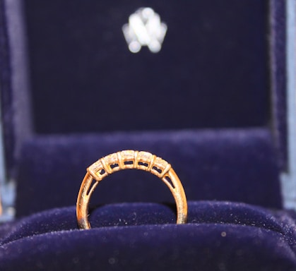 A gold ring with a row of small, sparkling diamonds is prominently displayed against a plush, dark blue velvet background. The ring appears elegant and is encased in a box lined with the same blue velvet, suggesting it is a piece of fine jewelry.