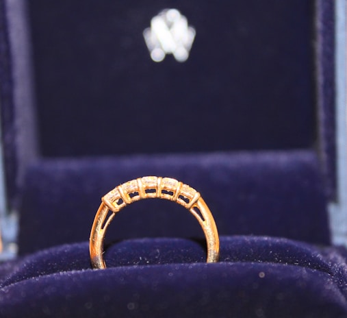 A gold ring with a row of small, sparkling diamonds is prominently displayed against a plush, dark blue velvet background. The ring appears elegant and is encased in a box lined with the same blue velvet, suggesting it is a piece of fine jewelry.