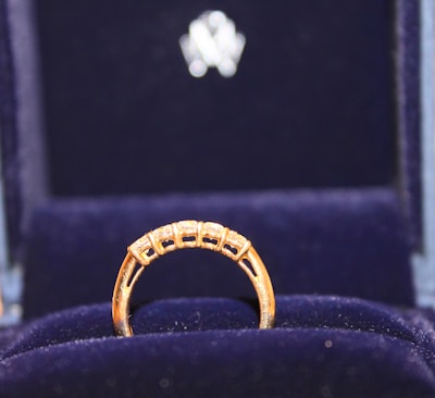 A gold ring with a row of small, sparkling diamonds is prominently displayed against a plush, dark blue velvet background. The ring appears elegant and is encased in a box lined with the same blue velvet, suggesting it is a piece of fine jewelry.