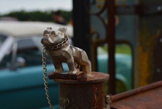 A metal bulldog figurine is secured with a chain on a rusty surface, potentially serving as a hood ornament. In the background, an out-of-focus vintage car with a soft blue-green color is visible, along with a grassy area and some indistinct objects.