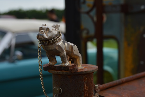 A metal bulldog figurine is secured with a chain on a rusty surface, potentially serving as a hood ornament. In the background, an out-of-focus vintage car with a soft blue-green color is visible, along with a grassy area and some indistinct objects.