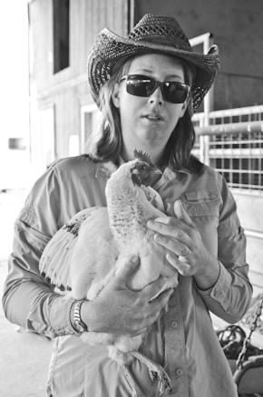 A person wearing a cowboy hat and sunglasses holds a chicken in their arms, standing in what appears to be a barn with metal fencing visible in the background. The black and white photograph captures a casual moment.