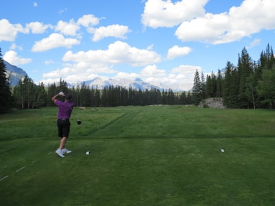 A golfer wearing a purple shirt and black shorts is captured mid-swing on a golf course surrounded by lush greenery. In the background, there are towering pine trees and a range of mountains under a partly cloudy blue sky. The teeing ground is well-maintained with neatly cut grass.