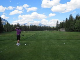 A golfer wearing a purple shirt and black shorts is captured mid-swing on a golf course surrounded by lush greenery. In the background, there are towering pine trees and a range of mountains under a partly cloudy blue sky. The teeing ground is well-maintained with neatly cut grass.