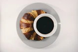 Freshly brewed coffee in a simple ceramic cup next to a croissant on a white marble countertop.