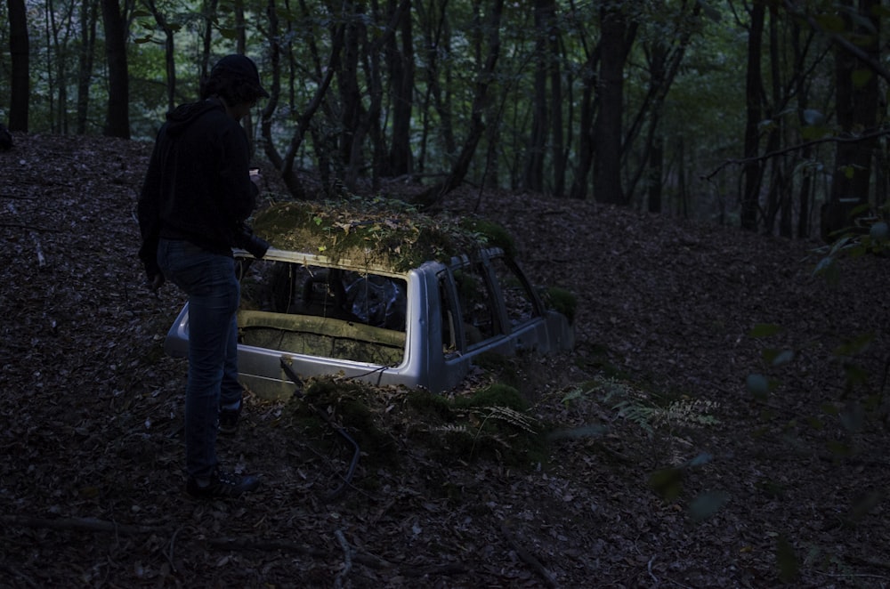 Fotografía de paisaje de hombre de pie frente a un coche roto medio enterrado en el bosque