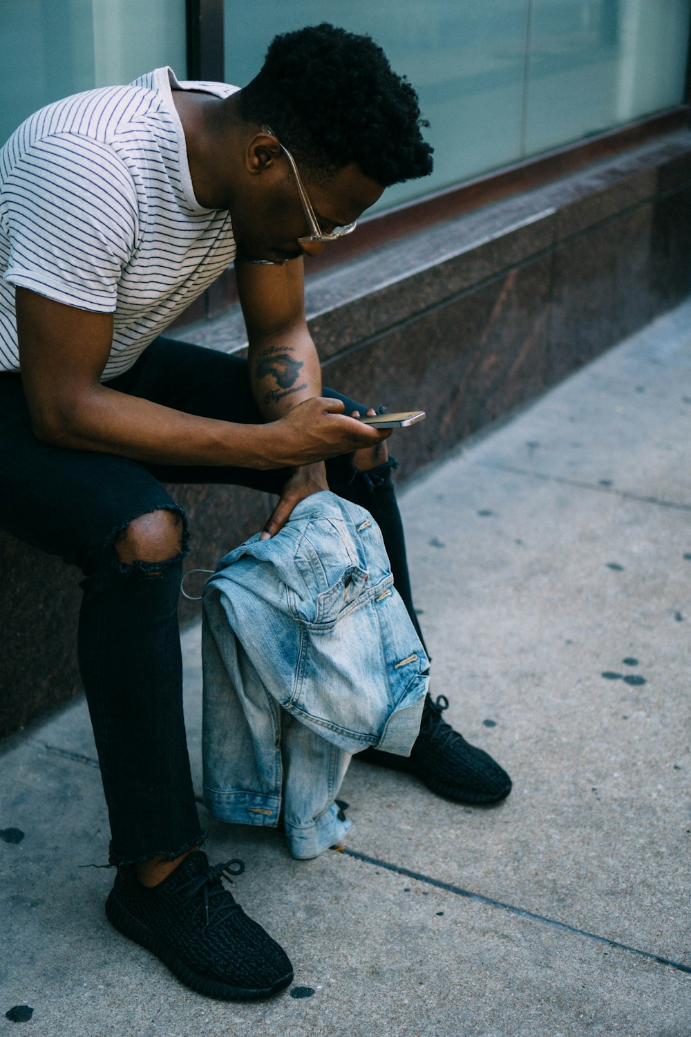 A man wearing a striped shirt and ripped jeans, sitting on a ledge, playing with his cellphone