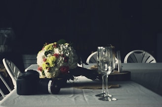 A dimly lit table setting featuring a bouquet of flowers with white and red blooms, a DSLR camera with a lens, and two empty wine glasses. The table is covered with a white cloth and surrounded by white folding chairs.