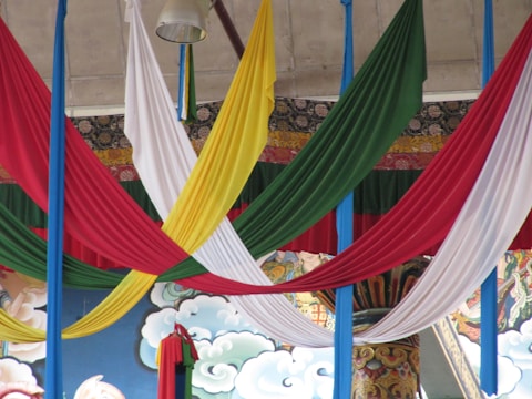 Brightly colored fabric drapes in red, green, yellow, and white hang from the ceiling, creating a vibrant and festive appearance. The background features artistic motifs with detailed patterns and decorative elements such as clouds and traditional designs.