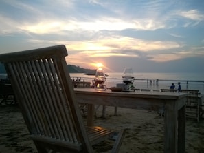 A serene beach setting during sunset, where wooden tables and chairs are positioned on the sand. The sun is low on the horizon, casting warm orange and pink hues across the sky and reflecting on the calm ocean waters. In the foreground, lanterns are placed on the table, and in the background, small groups of people are visible near the waterline.