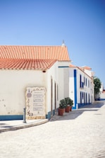 A sunlit street in a quaint village features white buildings with blue accents and red-tiled roofs. A signpost with directional information is fixed near the corner of a building. Terra cotta pots with green plants line the cobblestone pathway, with a clear blue sky overhead.