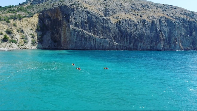 A tranquil coastal scene with vibrant turquoise water. Kayakers in brightly colored kayaks navigate the calm sea. A large, rugged cliff looms in the background, its rocky surface dominating the horizon.