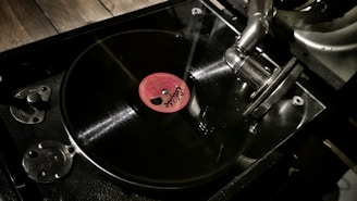 A close-up view of a vintage gramophone with a black vinyl record spinning on its turntable. The gramophone features a metallic arm and needle, with a prominent red label at the center of the record. The background includes a wooden surface, suggesting a classic or nostalgic setting.