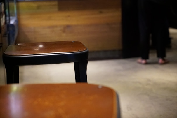 Two wooden bar stools with metal legs are in the foreground. The setting appears to be indoors with a wooden wall and a concrete floor. A person is visible in the background, standing and wearing dark clothing with sandals.
