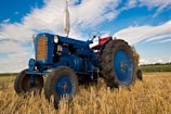 An old blue tractor is parked in a harvested field with straw on the ground. The sky is partly cloudy, and the fields extend into the distance. The tractor has large rubber tires and metal parts that show some wear, indicating regular use.