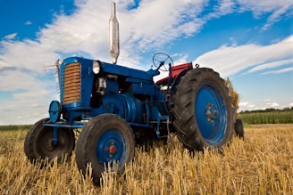 An old blue tractor is parked in a harvested field with straw on the ground. The sky is partly cloudy, and the fields extend into the distance. The tractor has large rubber tires and metal parts that show some wear, indicating regular use.