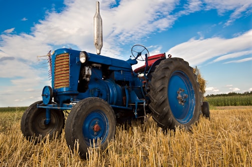 An old blue tractor is parked in a harvested field with straw on the ground. The sky is partly cloudy, and the fields extend into the distance. The tractor has large rubber tires and metal parts that show some wear, indicating regular use.