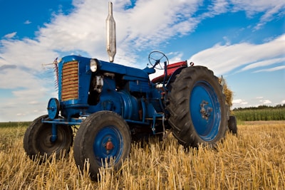 An old blue tractor is parked in a harvested field with straw on the ground. The sky is partly cloudy, and the fields extend into the distance. The tractor has large rubber tires and metal parts that show some wear, indicating regular use.