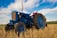 An old blue tractor is parked in a harvested field with straw on the ground. The sky is partly cloudy, and the fields extend into the distance. The tractor has large rubber tires and metal parts that show some wear, indicating regular use.