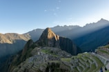 Machu Picchu is set against a backdrop of towering green mountains with beams of sunlight filtering through. The ancient Incan ruins are visible with their stone structures and terraces. A clear blue sky stretches above and tourists can be seen walking along the pathways.