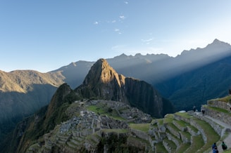 Machu Picchu is set against a backdrop of towering green mountains with beams of sunlight filtering through. The ancient Incan ruins are visible with their stone structures and terraces. A clear blue sky stretches above and tourists can be seen walking along the pathways.