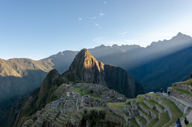 Machu Picchu is set against a backdrop of towering green mountains with beams of sunlight filtering through. The ancient Incan ruins are visible with their stone structures and terraces. A clear blue sky stretches above and tourists can be seen walking along the pathways.