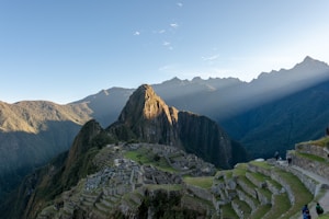 Machu Picchu is set against a backdrop of towering green mountains with beams of sunlight filtering through. The ancient Incan ruins are visible with their stone structures and terraces. A clear blue sky stretches above and tourists can be seen walking along the pathways.