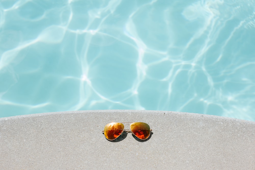 occhiali da sole in stile aviatore con lenti arancioni con montatura dorata accanto alla piscina