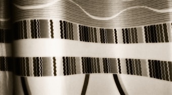 A close-up view of a patterned fabric with various horizontal stripes and wavy designs. The intricate patterns include a mix of thin and thick lines, and the overall color scheme appears muted with a sepia tone.