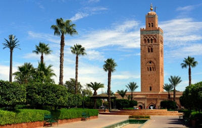 A tall, ornate minaret stands prominently against a bright blue sky with wispy clouds. Surrounding the structure are several tall palm trees and well-maintained gardens featuring lush green hedges and pathways. The architecture of the tower is intricate, with detailed carvings and geometric patterns typical of traditional Moroccan design.