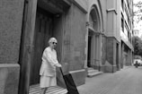 An elderly woman wearing sunglasses stands on a tiled doorstep, holding onto a small trolley. She is positioned beside a large, ornate entrance with detailed brickwork and an arched façade. The scene is set on a quiet city street lined with modern buildings.