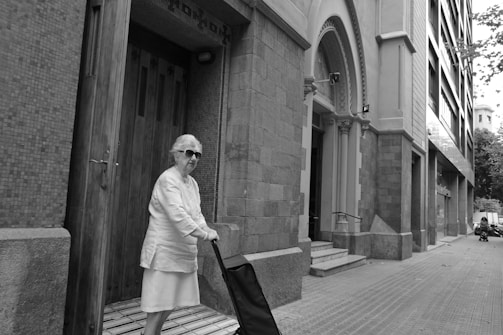 An elderly woman wearing sunglasses stands on a tiled doorstep, holding onto a small trolley. She is positioned beside a large, ornate entrance with detailed brickwork and an arched façade. The scene is set on a quiet city street lined with modern buildings.