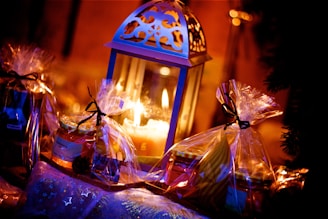 A warmly lit scene featuring a decorative lantern with intricate designs, surrounded by gift packages wrapped in clear plastic with black bows. The setting is cozy and festive, with a glowing candle inside the lantern casting a soft, ambient light, highlighting the holiday spirit.