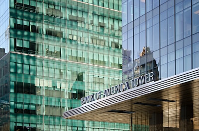 A modern, glass-fronted skyscraper with reflective windows. The words 'Bank of America Tower' are prominently displayed on the building. The structure features sleek lines and a contemporary architectural style.
