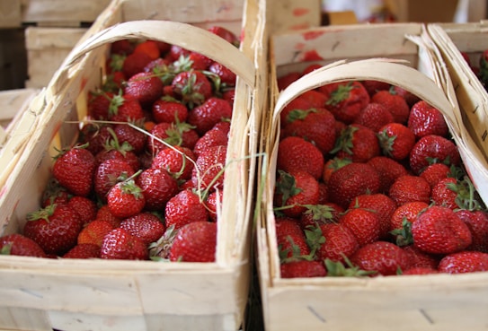 Wooden baskets overflowing with fresh, ripe strawberries are arranged closely together. The strawberries are vibrant red, with some green leaves still attached. The baskets are rustic, made from light-colored wood.