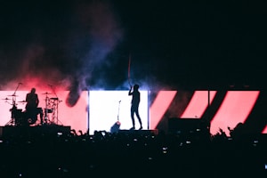 A silhouetted performance on stage with vibrant lighting effects in the background, displaying dramatic reds, blues, and white. A drummer is positioned to the left, while a singer or performer stands to the right, facing the audience. Smoke and haze add to the dynamic atmosphere.