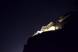 A modern house with prominent geometric architecture is perched on a hillside, illuminated against a dark night sky. The structure features multiple levels and angular lines, with bright lights casting stark shadows.