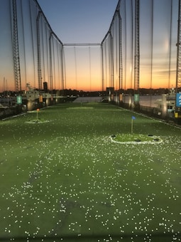 A golf driving range at sunset, with a field filled with scattered golf balls. The sky is painted in shades of orange and blue as the sun sets on the horizon. Tall netted fences surround the area, and there are a few targets marked by flags on the ground. The waterfront can be seen in the distance with some boats docked along the shore.