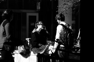 A black and white photograph capturing three people interacting in an urban setting. One person with a backpack stands facing another who is seated on a ledge, wearing sunglasses and a polka dot dress. A third person is walking past them carrying a bag, partially obscured. The scene is set outdoors with a mix of natural and artificial light casting strong contrasts.